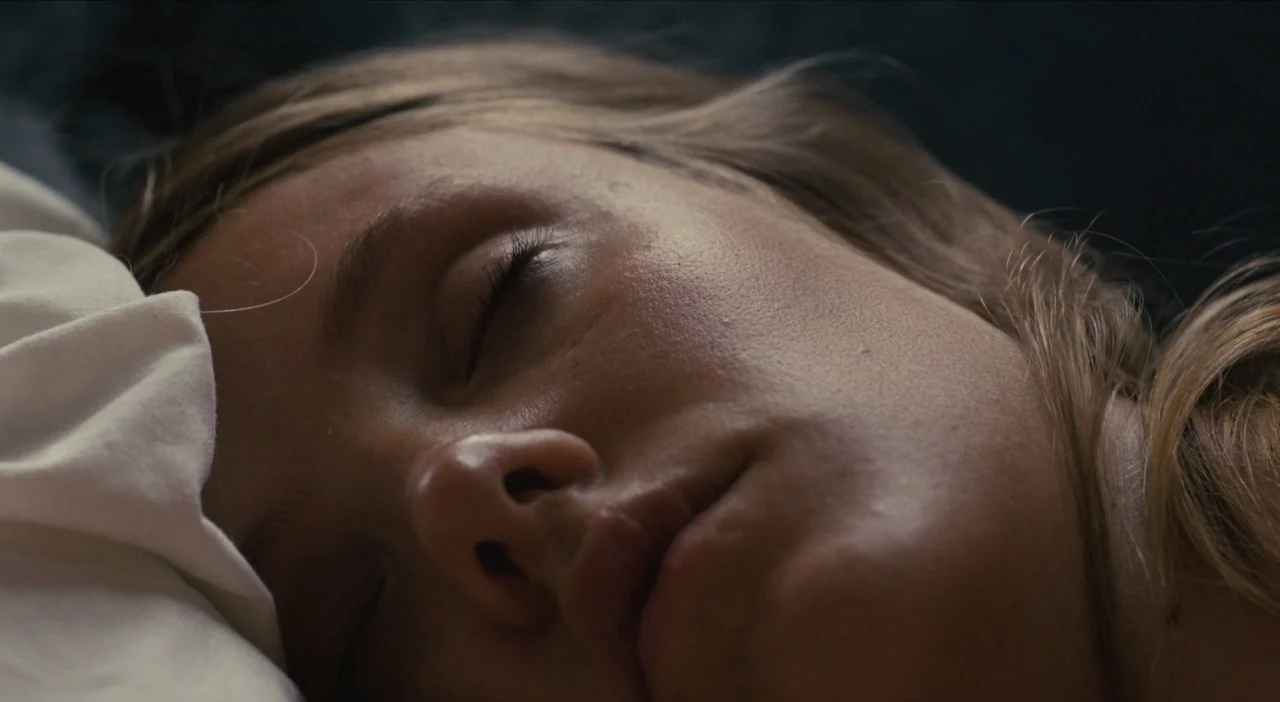 The Comedy (2012) movie still: closeup — A close view shows a face resting on a white pillow in dim light. The woman lies still with her eyes closed.