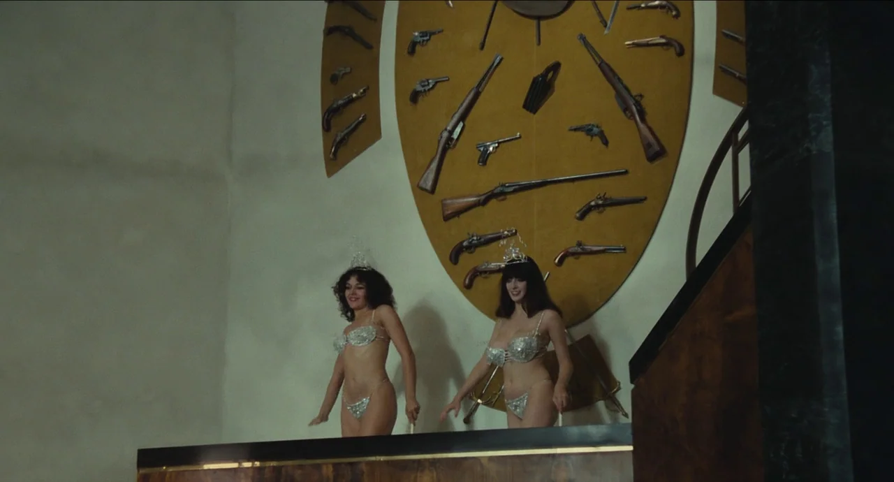 City of Women (1980) movie still: fullbody — Two women stand on a stage before an oval-shaped wall mount displaying antique firearms. They wear…