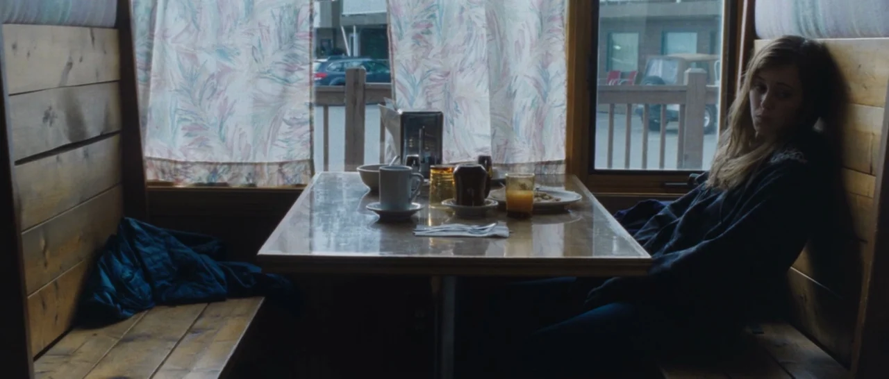 Wildlike (2014) movie still: medium — A wooden booth in a diner features patterned curtains over a large window. A woman sits alone at the table…