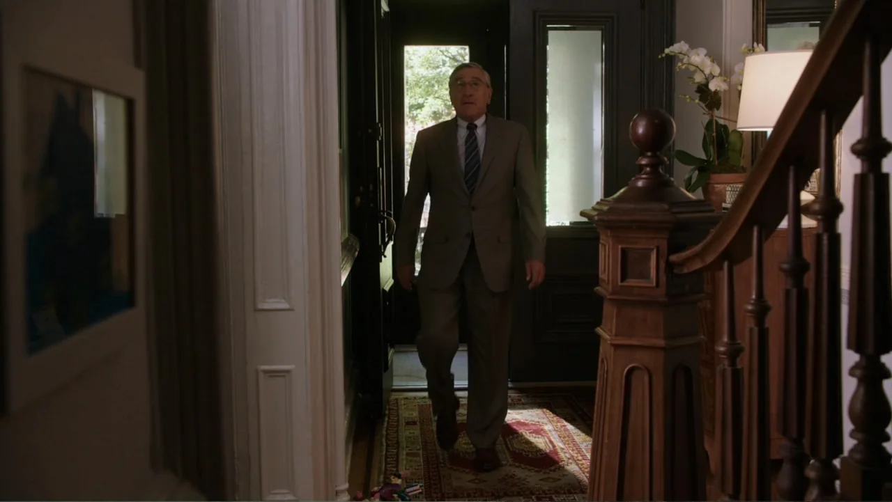 The Intern (2015) movie still: fullbody — An entryway features a dark wooden staircase and an ornate rug on the floor. A man in a suit enters the…