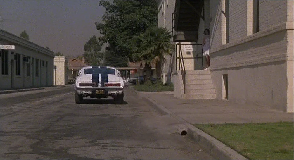 Coming Home (1978) movie still: wide — A quiet road features a white sports car driving away from the camera. A person in a white dress stands on a…