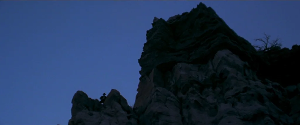 In A Valley of Violence (2016) movie still: wide — A dark, craggy rock formation rises against a clear deep-blue twilight sky. A single small human…