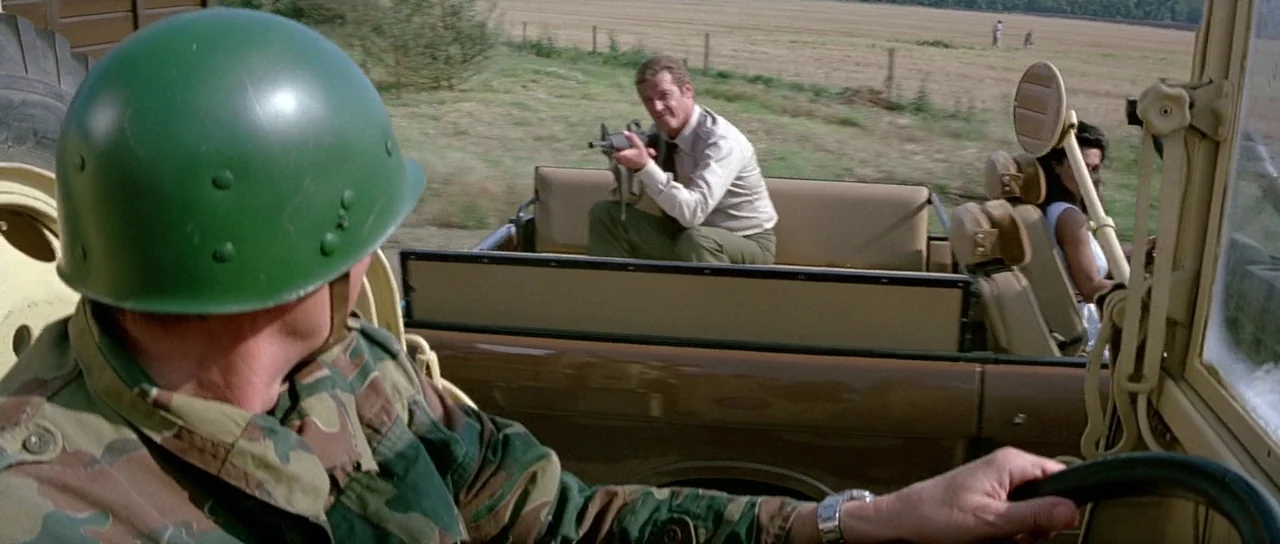 Octopussy (1983) movie still: medium — A field with a dirt track where a brown jeep drives forward. A man with a rifle sits in the rear passenger…