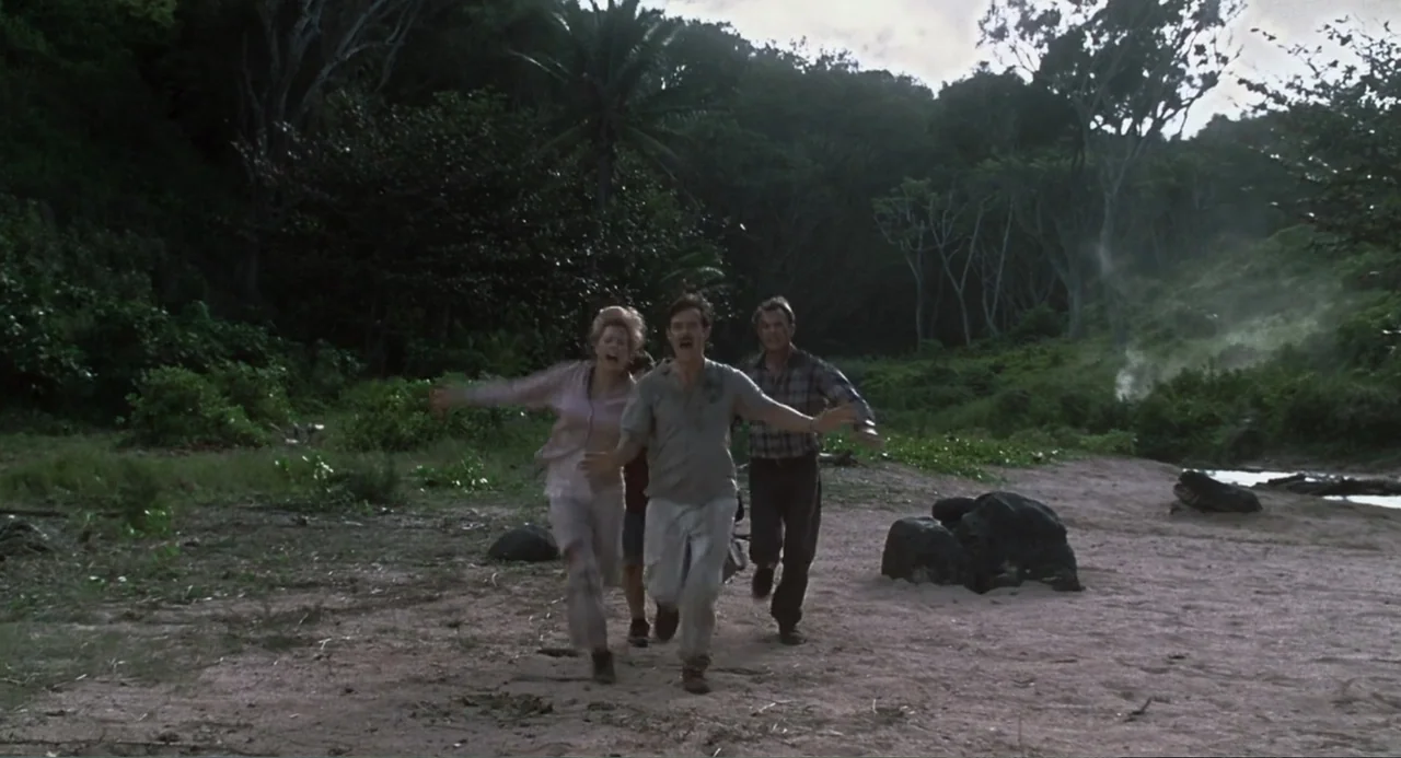 Jurassic Park III (2001) movie still: fullbody — A remote sandy beach borders a dense, dark tropical jungle with thick foliage. Three adults run…