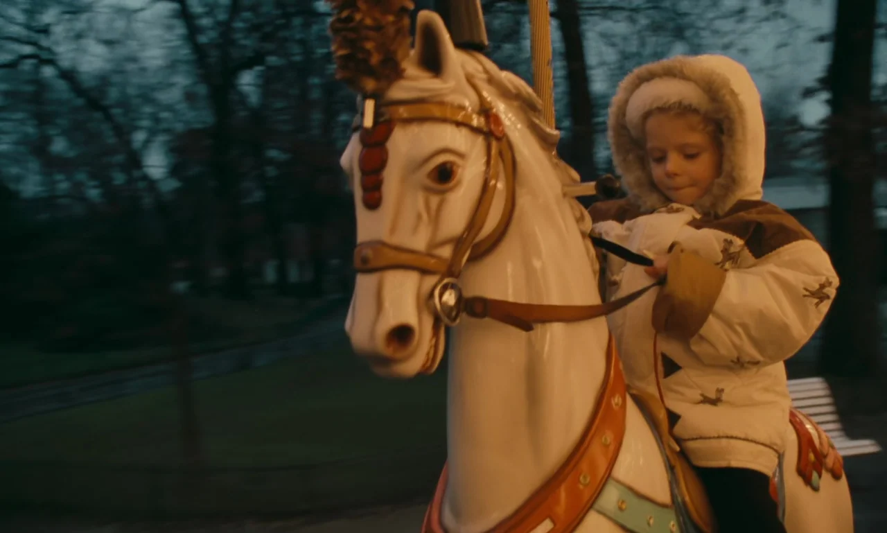 A Tale of Winter (1992) movie still: medium — An outdoor amusement park at dusk features a turning carousel ride with dark trees. A young child…