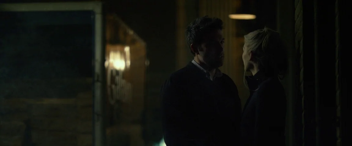 Gone Girl (2014) movie still: medium — A dark night street area with a blurred storefront sign visible in the background. A man and a woman stand…