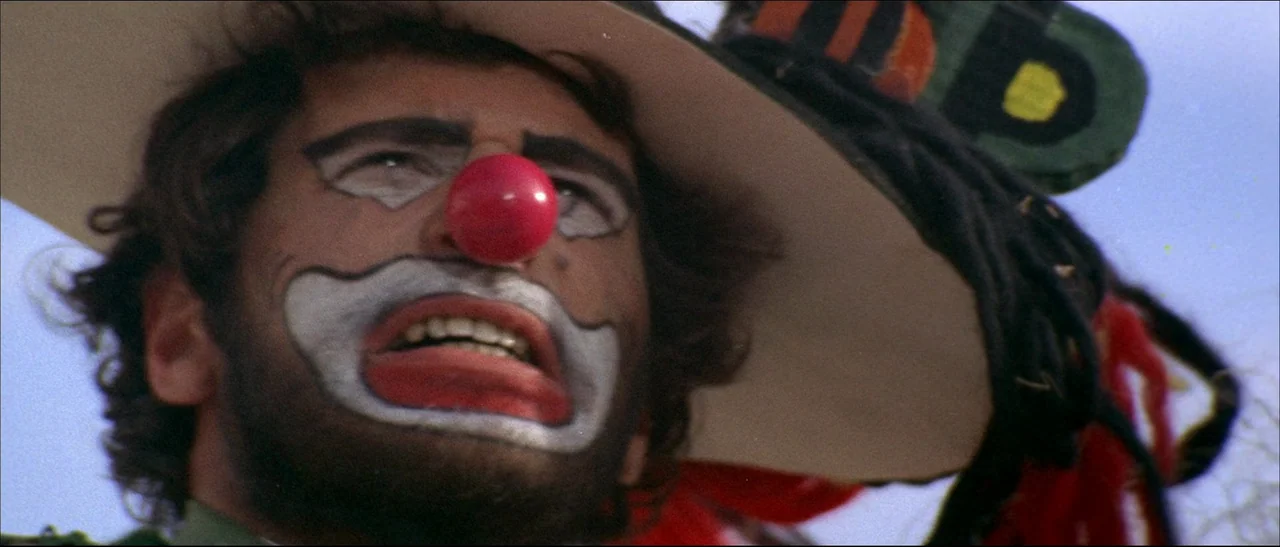 The Mercenary (1968) movie still: closeup — A man wearing clown makeup and a sombrero looks upward against a clear sky. He wears a red foam nose and…