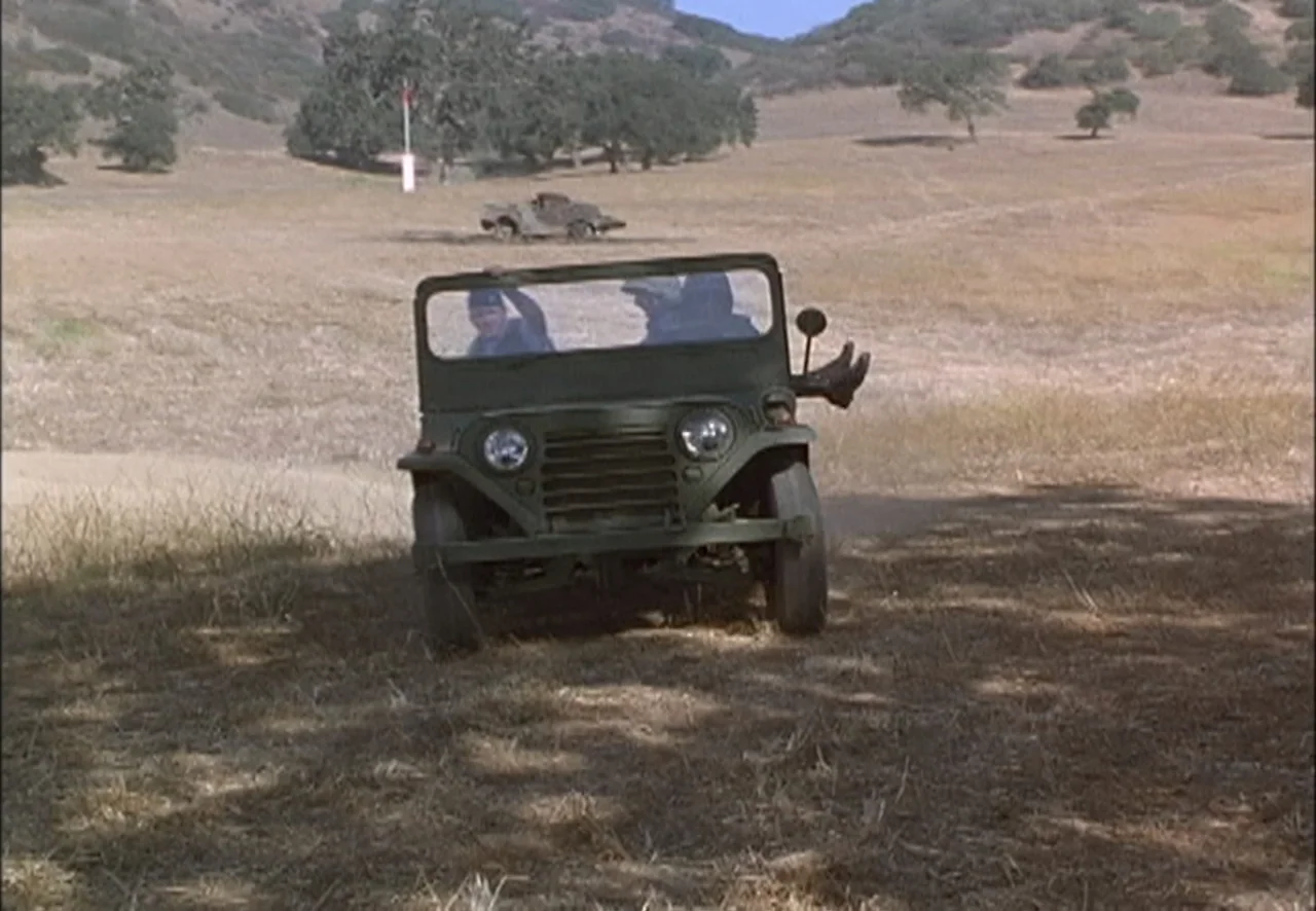 The Pentagon Wars (1998) movie still: medium — An olive-drab military jeep drives across a dry, grassy field with scattered oak trees. Two soldiers…