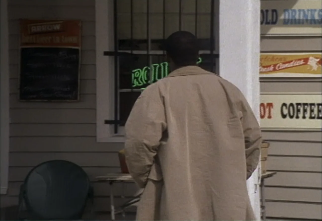 Ambushed (1998) movie still: medium — A storefront exterior features wooden siding with a menu board and various signs. A man wearing a tan coat…