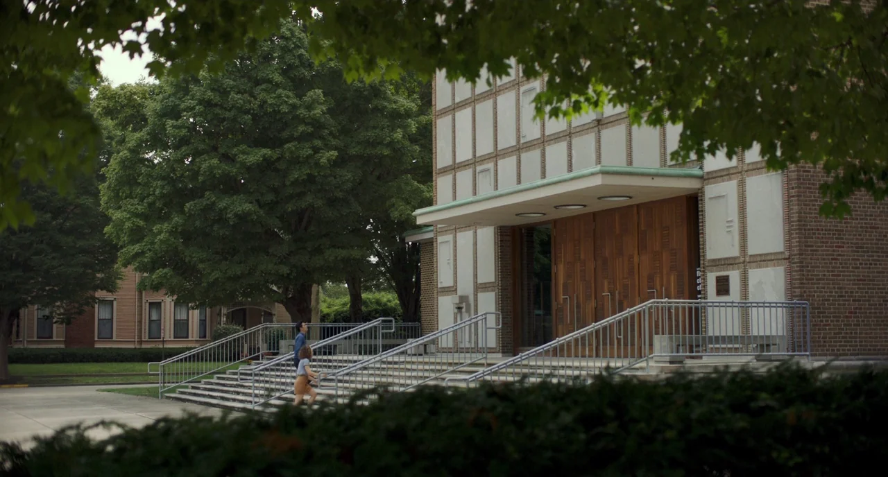 Columbus (2017) movie still: wide — A modern building with a grid-pattern facade and wood doors stands beside lush trees. A man and woman walk up…