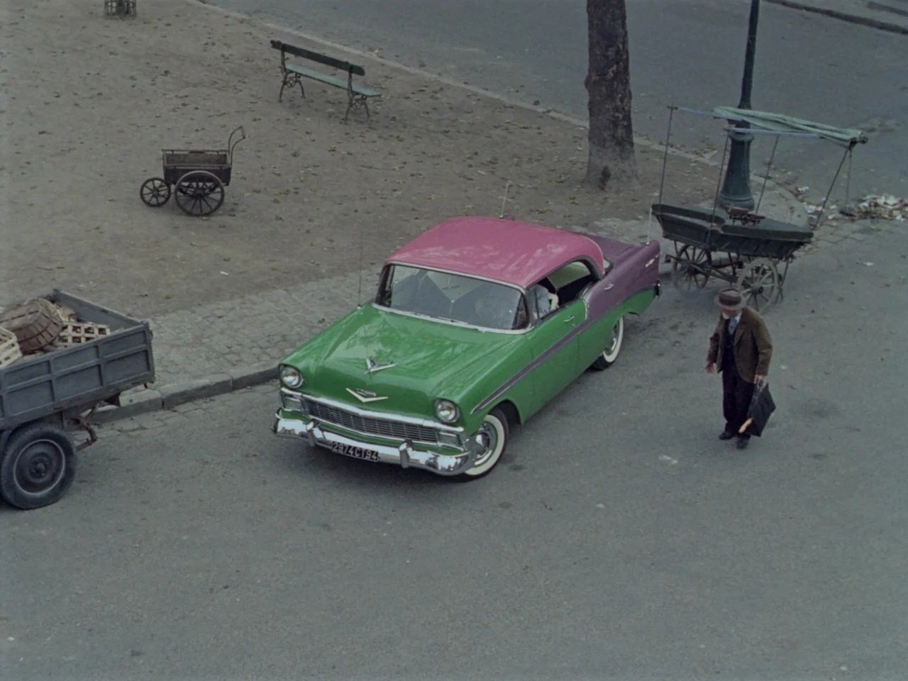 Mon Oncle (1958) movie still: wide — A paved city square features a vintage green and magenta car parked beside a cart. An elderly man in a brown…