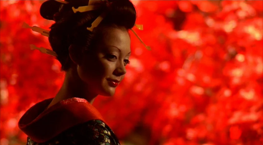 Sakuran (2006) movie still: closeup — A woman stands in front of a dense cluster of bright red autumn leaves. She is positioned in profile, framed…