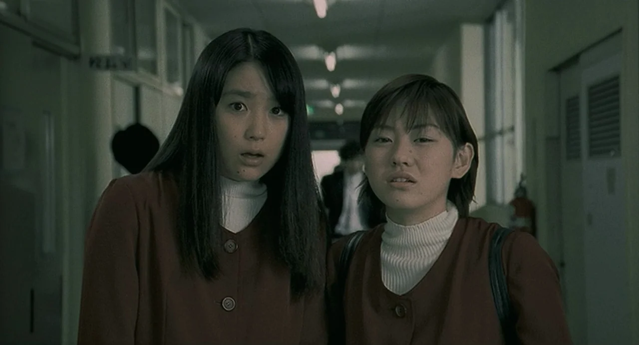 Uzumaki (2000) movie still: medium — A long school corridor with fluorescent lights and windows along the side wall. Two teenage girls stand in the…