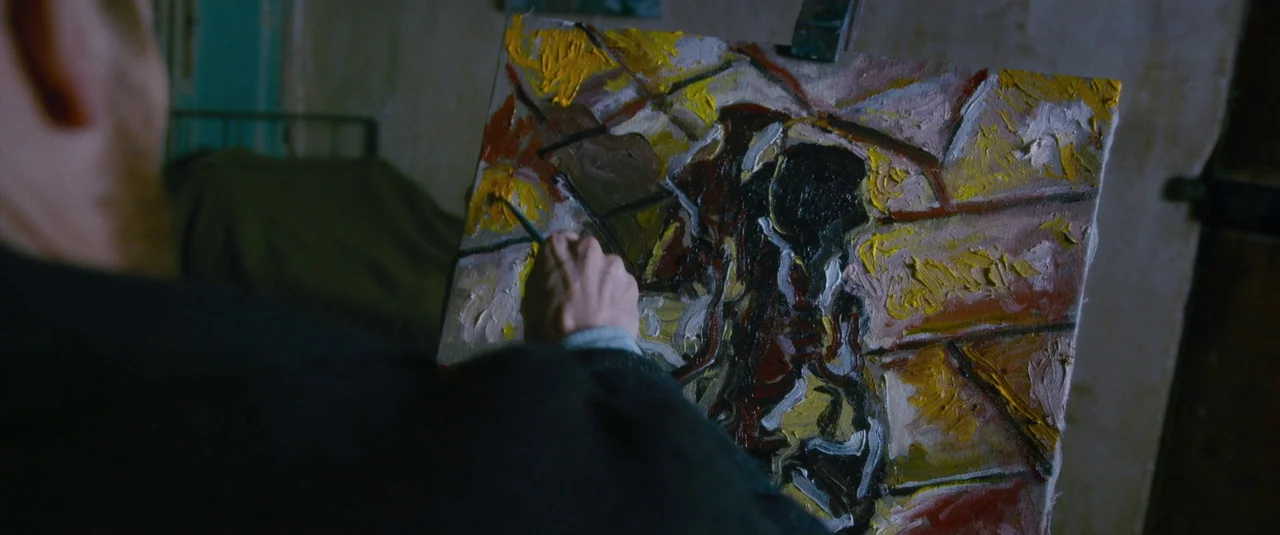 At Eternity’s Gate (2018) movie still: medium — A dimly lit room with an easel positioned in front of a neutral wall. A person paints on a canvas…
