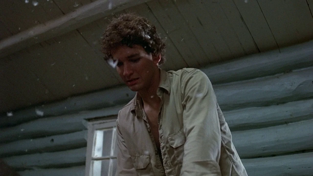 Friday the 13th (1980) movie still: medium — A rustic interior with stacked wooden log walls and a small rectangular window. A young man with curly…