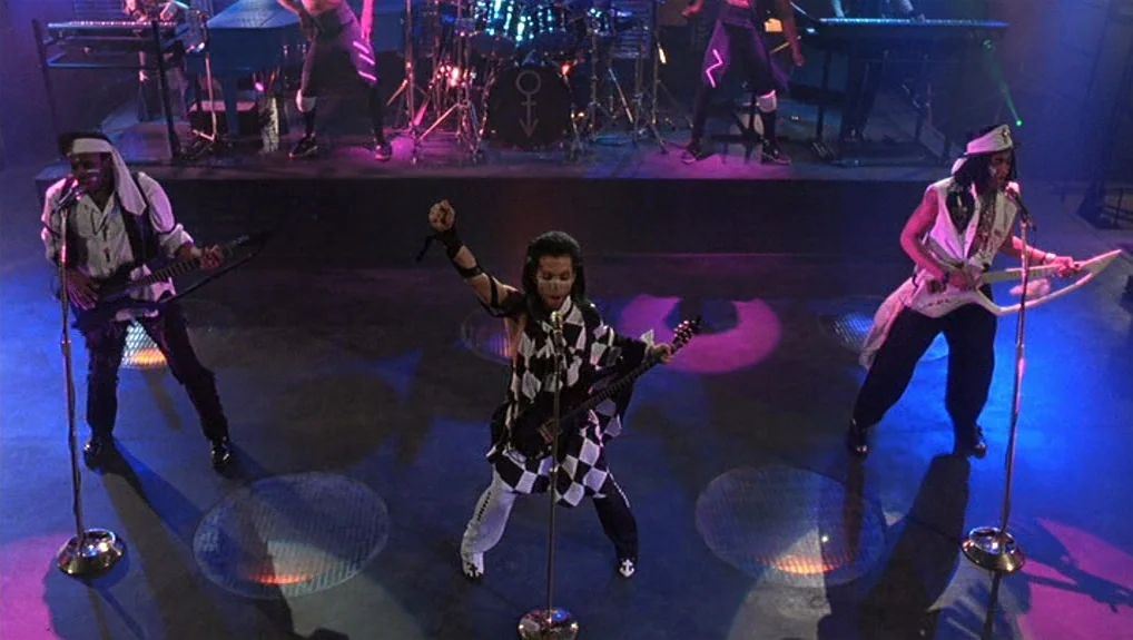Graffiti Bridge (1990) movie still: wide — A dark concert stage with musical instruments and performers bathed in vibrant colored spots. Prince…