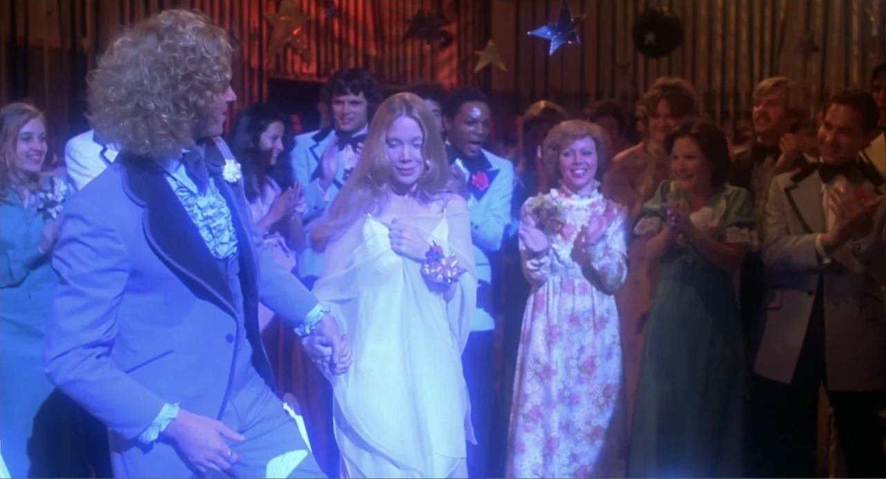 Carrie (1976) movie still: medium — A high school gym is decorated with hanging stars and streamers for a dance. A young couple walks hand-in-hand…