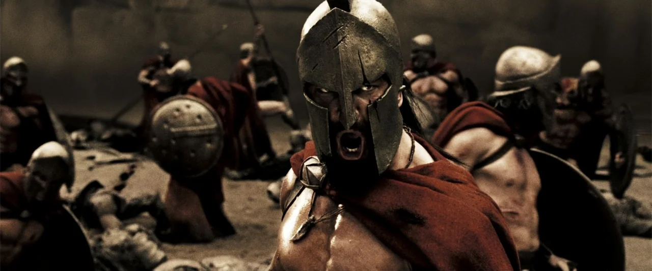 300 (2007) movie still: medium — A dusty, sun-drenched dirt ground serves as a battlefield for ancient warriors in combat. A muscular man in a…
