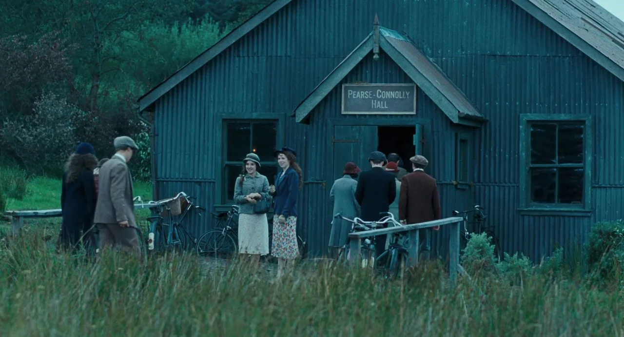 Jimmy’s Hall (2014) movie still: wide — A rural meeting hall made of corrugated metal sits in a grassy clearing. A group of people in 1920s attire…