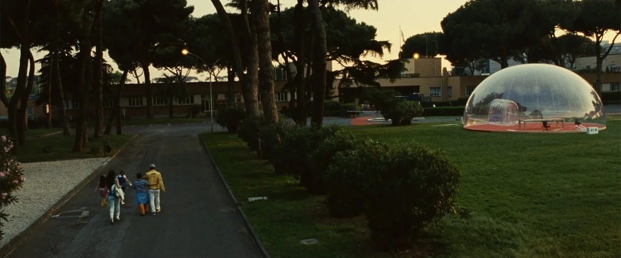 Reality (2023) movie still: wide — A group of people walks along a paved path lined with tall pine trees. A large transparent geodesic dome…