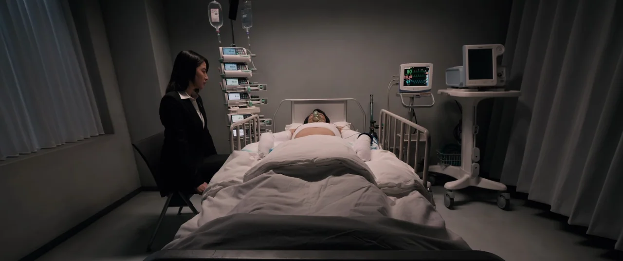 Shin Ultraman (2022) movie still: wide — A sterile medical room features a patient lying in a bed surrounded by equipment. A woman in a dark suit…