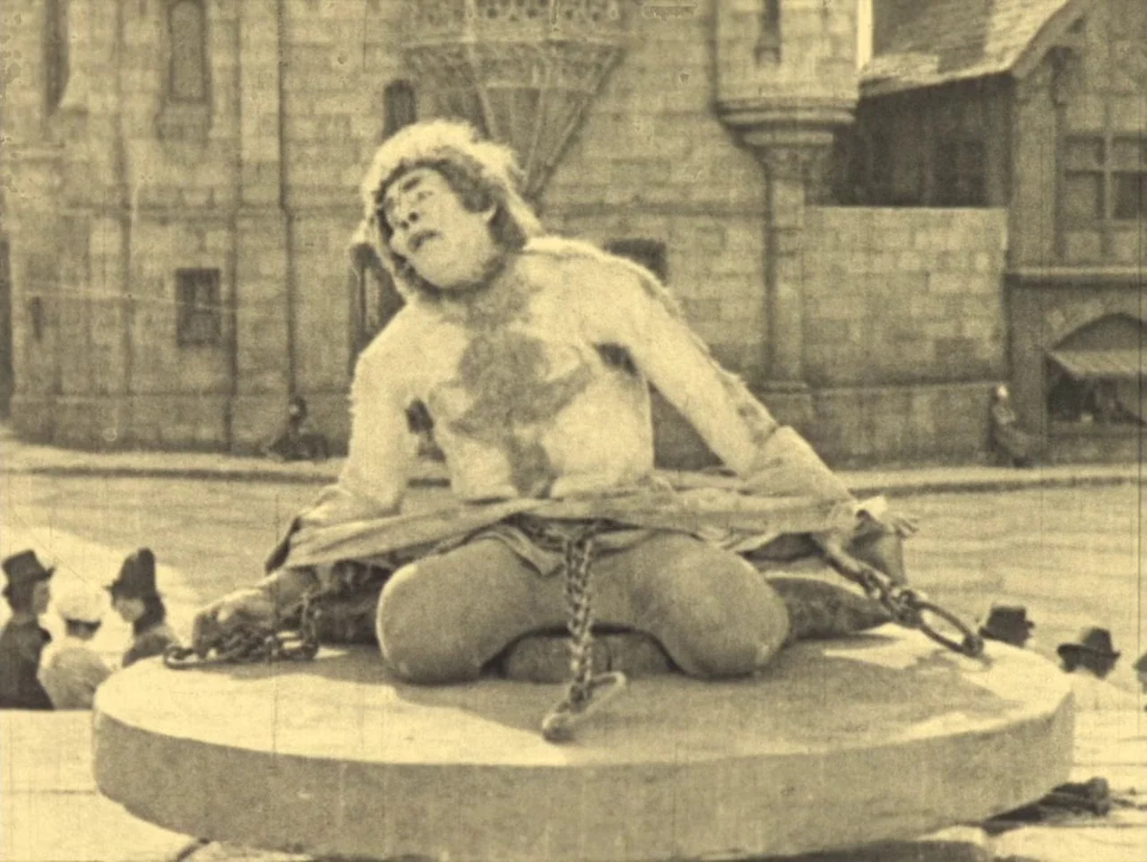 The Hunchback of Notre Dame (1923) movie still: fullbody — A man kneels on a stone platform in front of medieval-style stone buildings. He is…