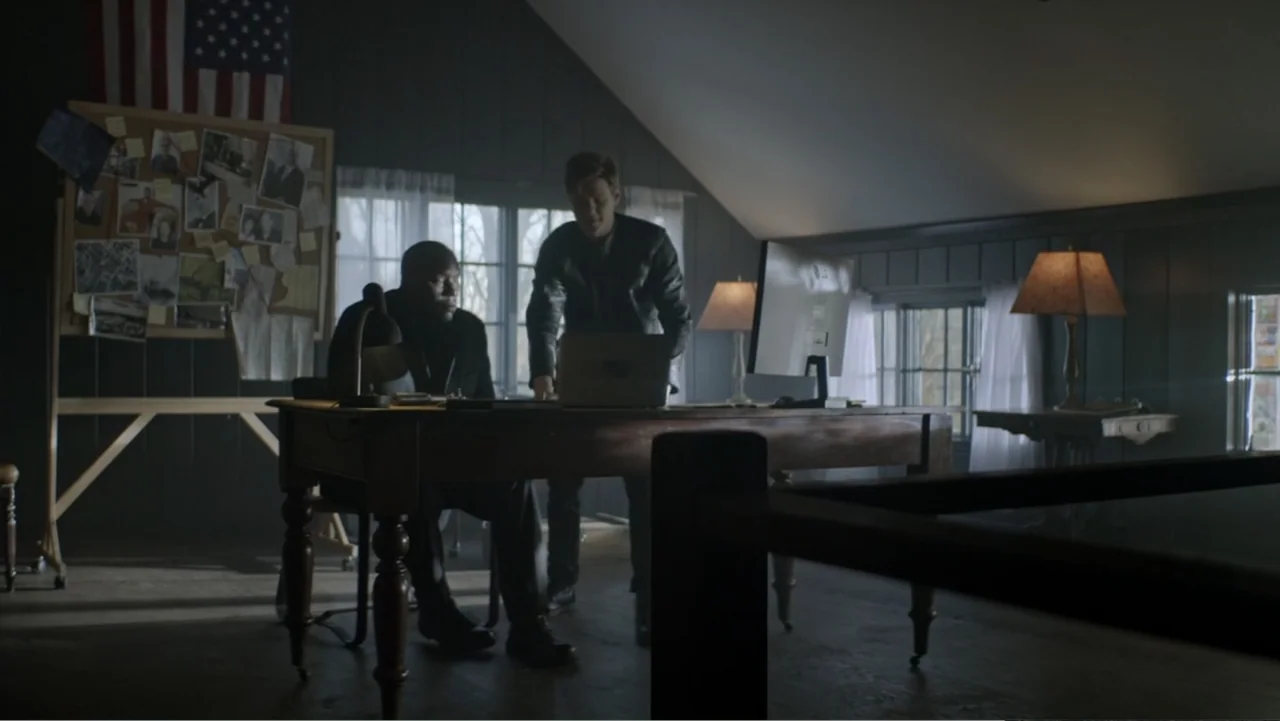 The Collective (2023) movie still: medium — A dimly lit attic converted into a workspace with dark paneling and sloped ceilings. Two men stand at a…