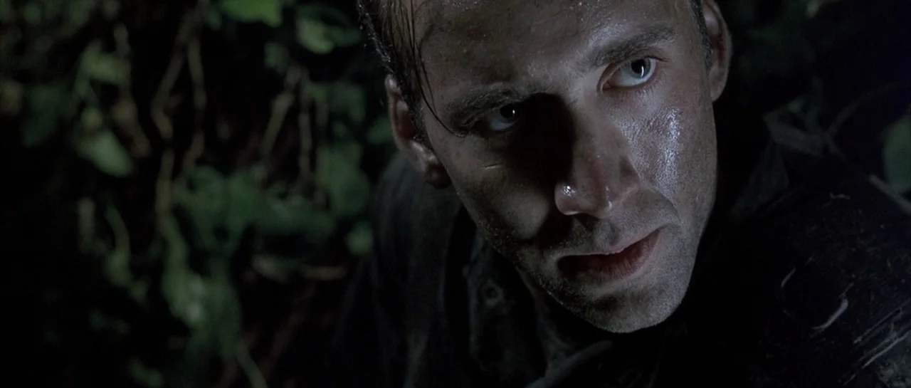 The Rock (1996) movie still: closeup — A man hides among dark, dense leaves at night under low-key lighting. The subject watches something…