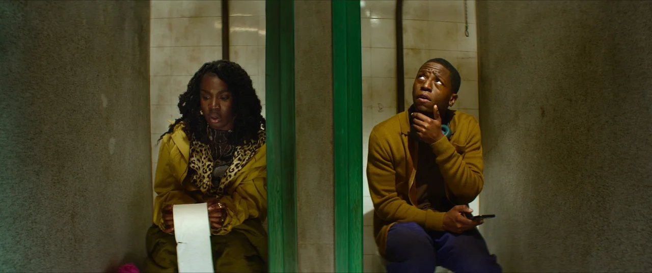 Rye Lane (2023) movie still: medium — Two individuals sit in adjacent public stalls separated by a green-painted wooden partition wall. The woman…