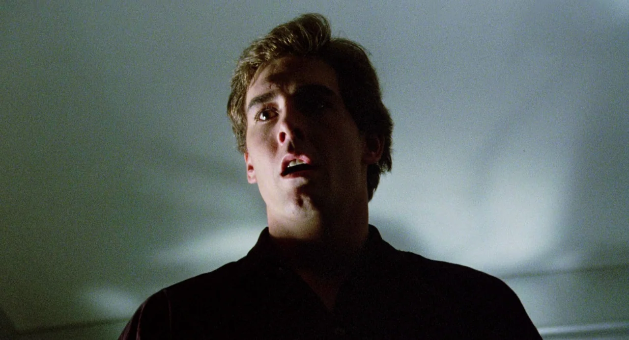 Crimes of Passion (1984) movie still: closeup — A dimly lit room provides a dark backdrop for the subject's face. A young man looks upward with a…