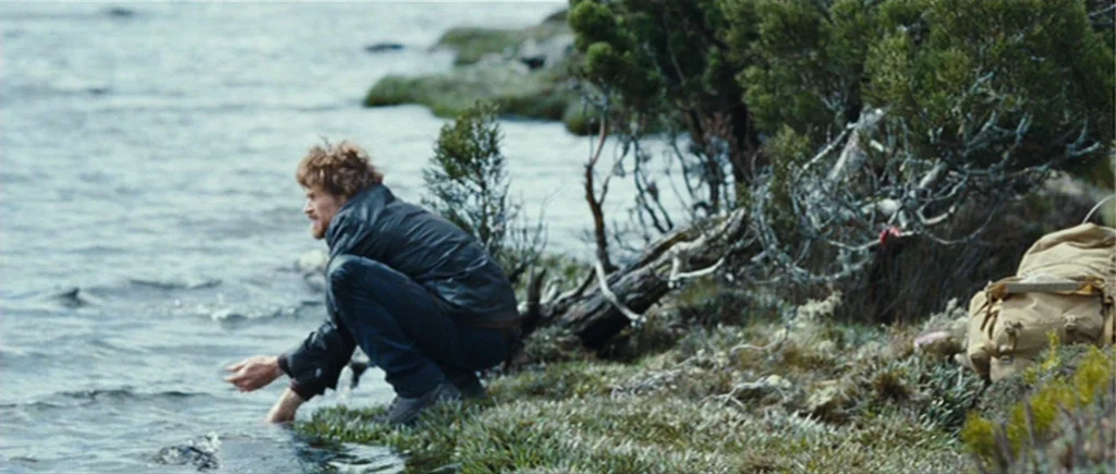 The Hunter (2011) movie still: medium — A rugged lakeside with sparse, low-lying shrubs and mossy ground on an overcast day. A man crouches at the…
