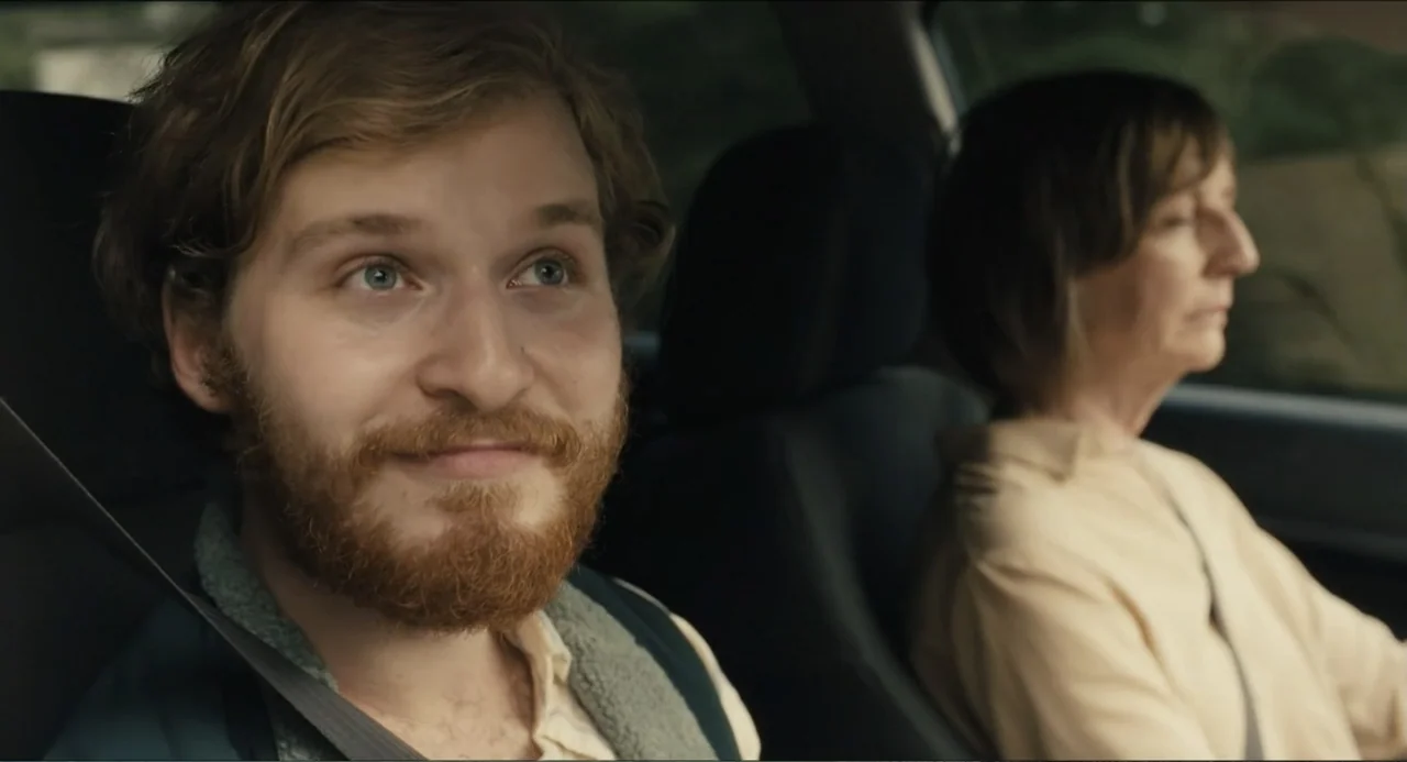 Hell of a Summer (2025) movie still: medium — Inside a moving vehicle, a man and an older woman are sitting side-by-side. The man faces slightly…
