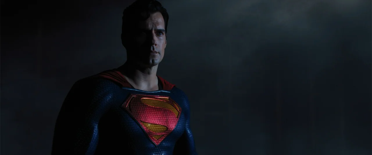 Black Adam (2022) movie still: medium — A dark, nondescript background frames the upper body of a man in a tactical suit. The subject remains static…