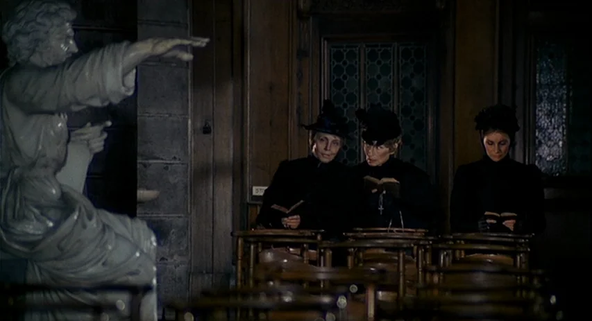 Malpertuis (1971) movie still: medium — Three women in black dresses and hats sit together in wooden pews reading books. A large white statue of a…