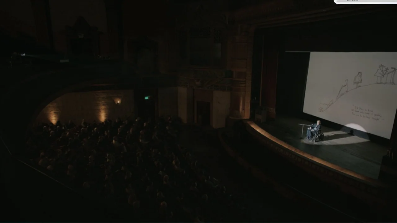 Don't Worry, He Won't Get Far on Foot (2018) movie still: wide — A dark auditorium features a solitary figure on a stage in front of a projection…