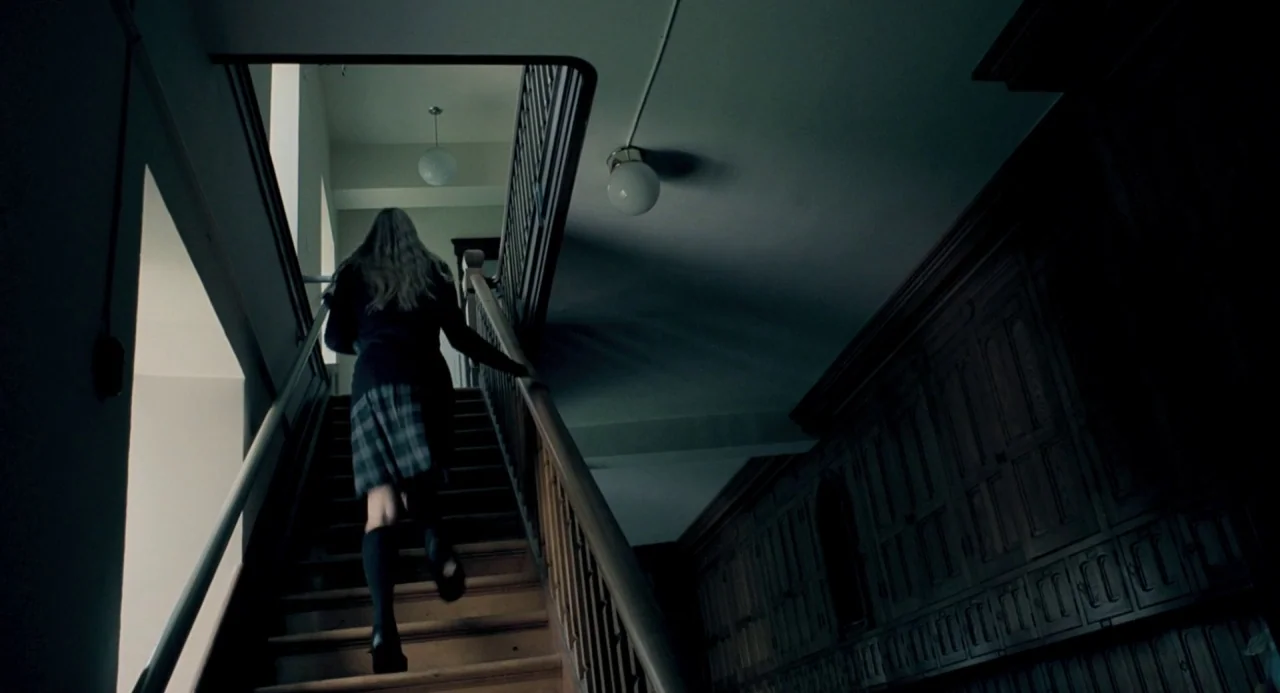 The Moth Diaries (2011) movie still: fullbody — A dimly lit wooden staircase features dark paneled walls and globe ceiling fixtures. A young person…