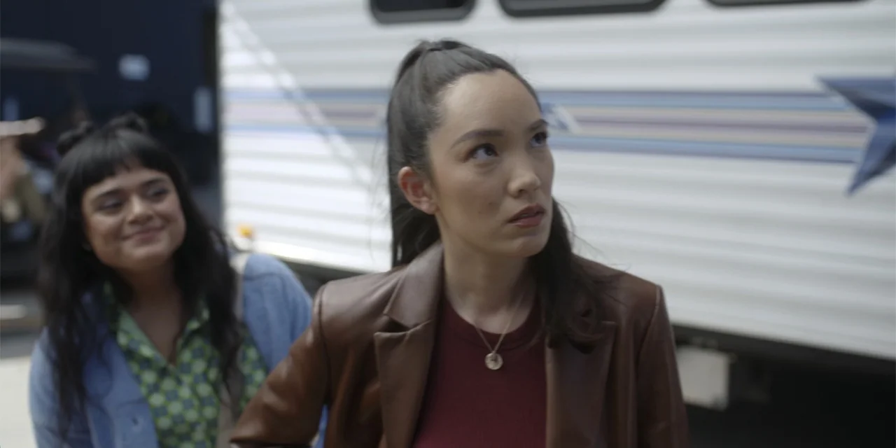 A Hollywood Christmas (2022) movie still: medium — A paved exterior area features a large white RV parked in the background. Two young women stand…