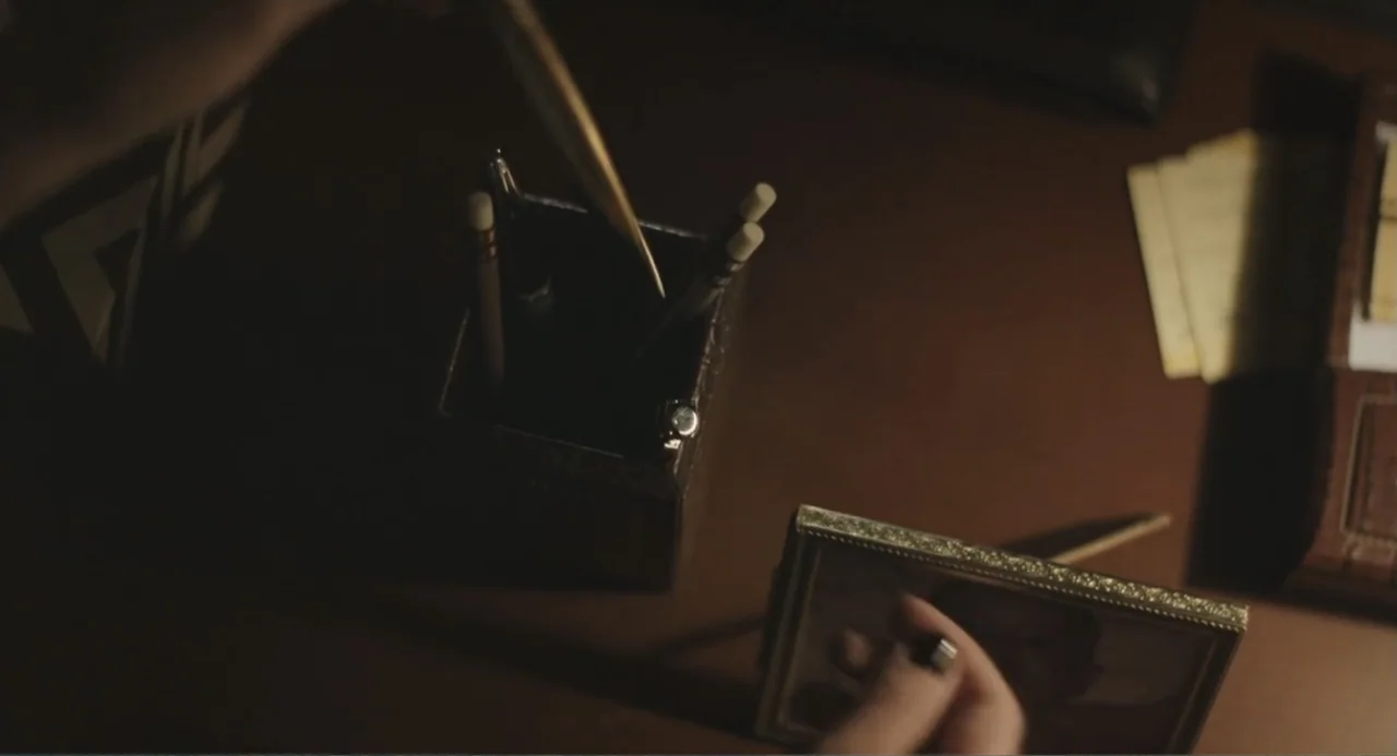 Beatriz at Dinner (2017) movie still: closeup — A dark wooden desk surface features a pencil cup and a small picture frame. A hand enters the frame…