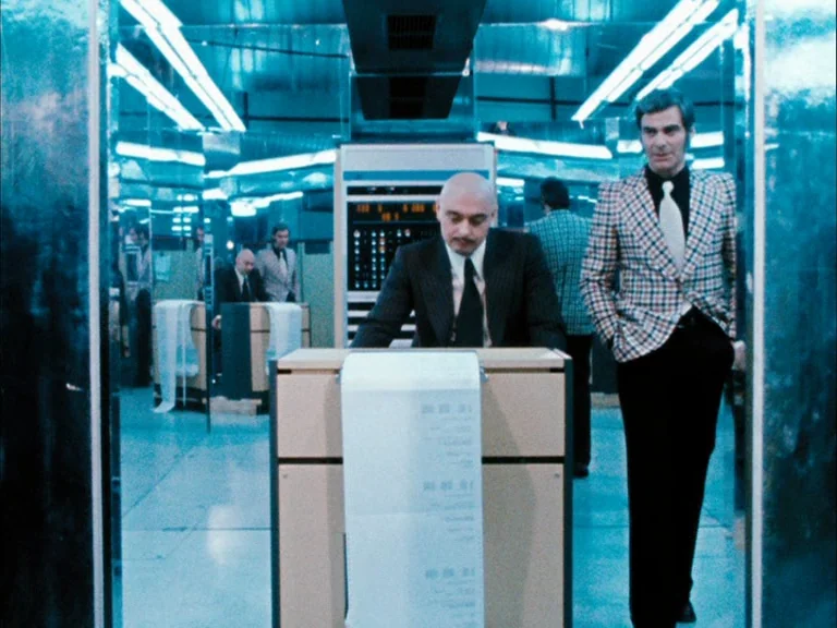 Making of: World On A Wire (1973) movie still: fullbody — A 1970s computer room is filled with mainframe hardware and rows of mirrored walls. A bald…
