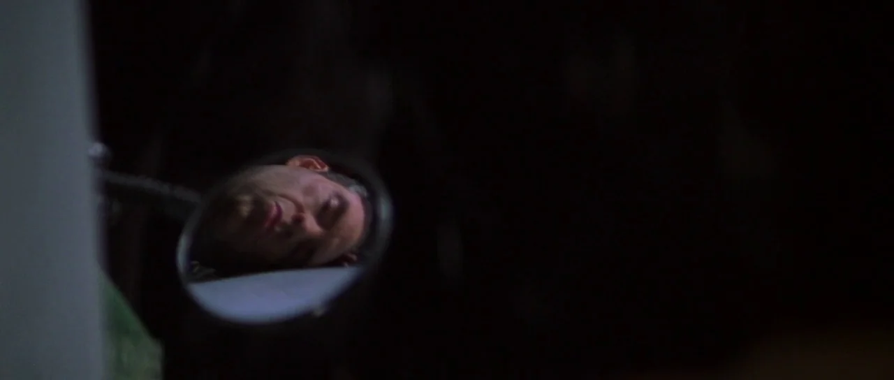 The Thomas Crown Affair (1999) movie still: closeup — A dark environment holds a small circular mirror reflecting a man's face. The man lies down…