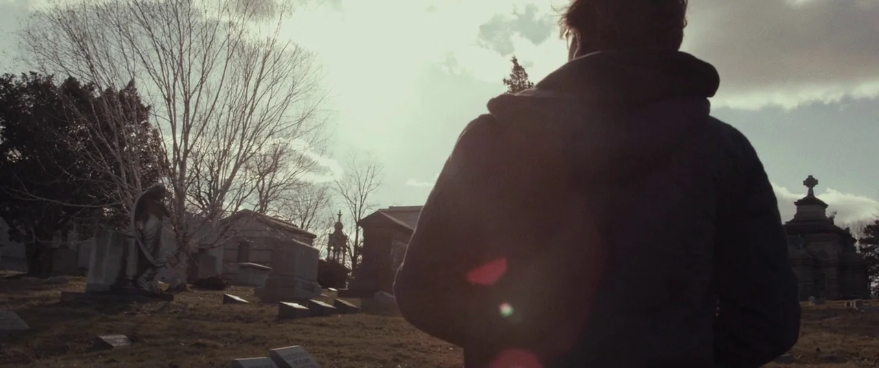 I Origins (2014) movie still: medium — A cemetery during the day with scattered gravestones and bare trees under clouds. A man in a dark hoodie…