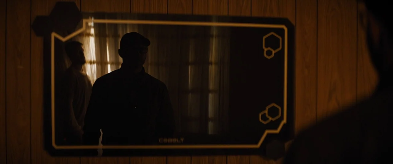 Upgrade (2018) movie still: medium — A wall covered in wood paneling features a rectangular black display surface. A man in a cap stands in front of…