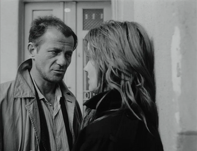 Damnation (1988) movie still: medium — A man and a woman stand close together in front of a closed wooden door. They look at each other while…