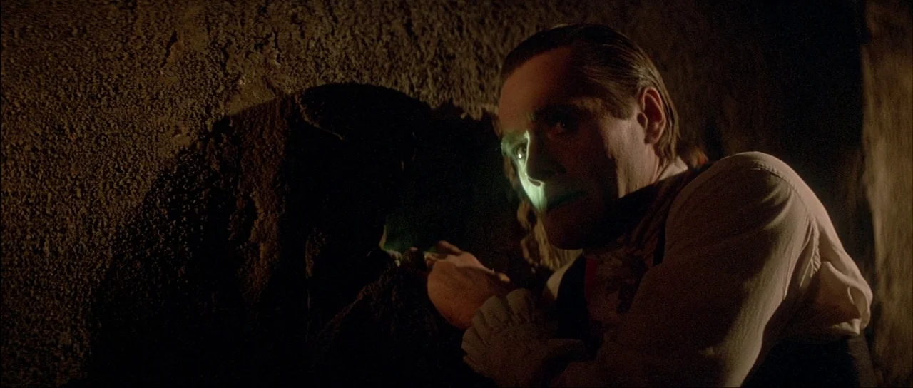 The Monster Squad (1987) movie still: medium — A dark, cavernous space is enclosed by rough-hewn stone walls. A man stares through a small hole in…