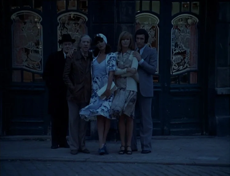 The Arrival of Joachim Stiller (1976) movie still: fullbody — A group of five people stands on a sidewalk in front of dark storefront doors. Each…
