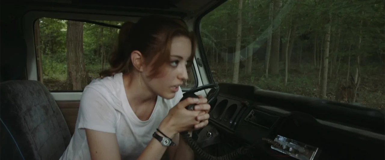Darkness Waits (2020) movie still: medium — The interior of a van parked in a forest during the daylight hours. A young woman holds a handheld radio…