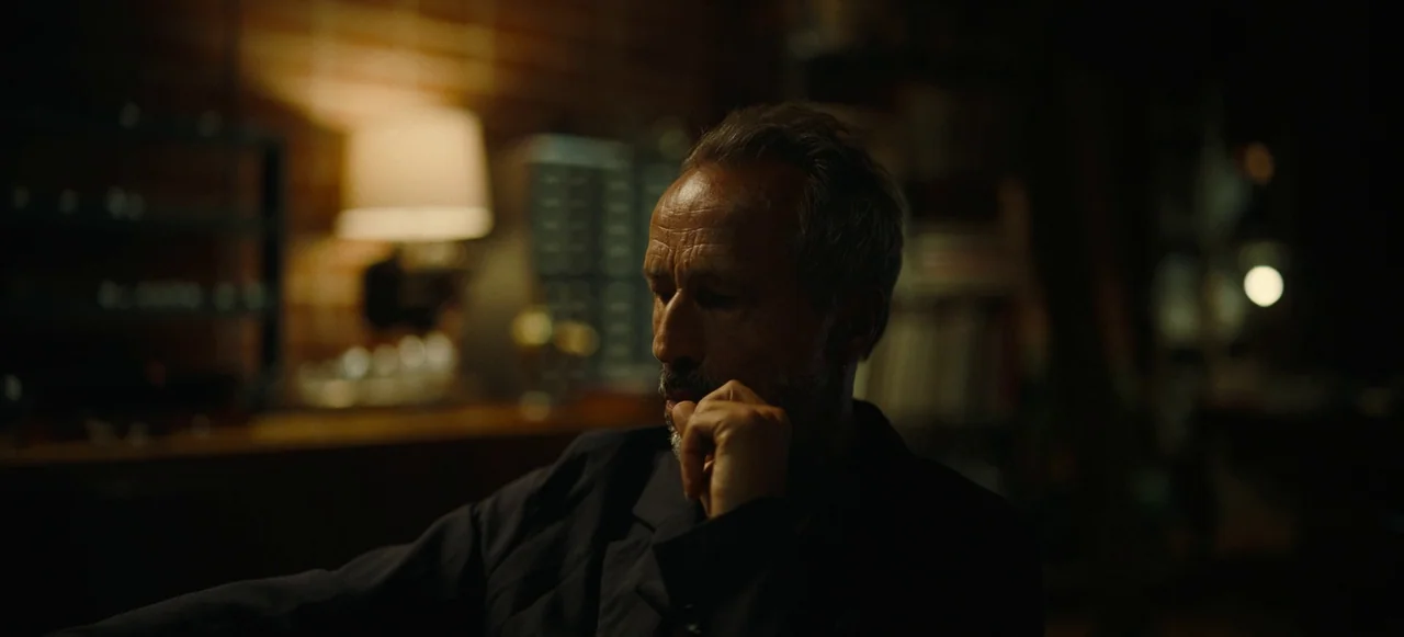 Nope (2022) movie still: medium — A dimly lit interior space features a brick wall background and a desk lamp. An older man with a gray beard sits…