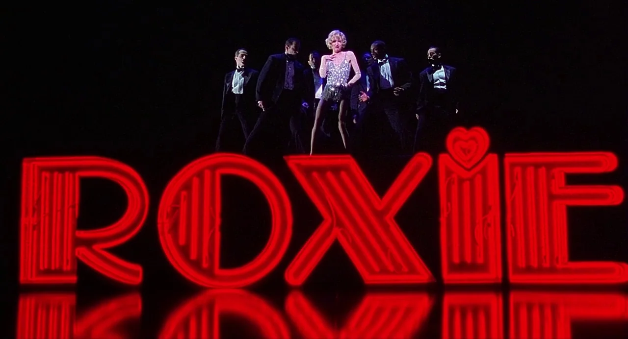 Chicago (2002) movie still: fullbody — A dark stage features a large red neon sign displaying the name ROXIE. A blonde woman and five men in tuxedos…