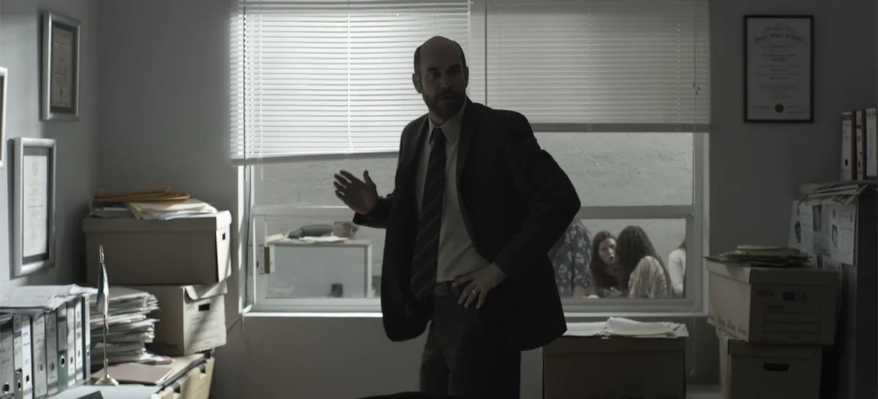 About the Deaths (2016) movie still: medium — A cluttered office features stacks of cardboard boxes, filing cabinets, and framed certificates on…