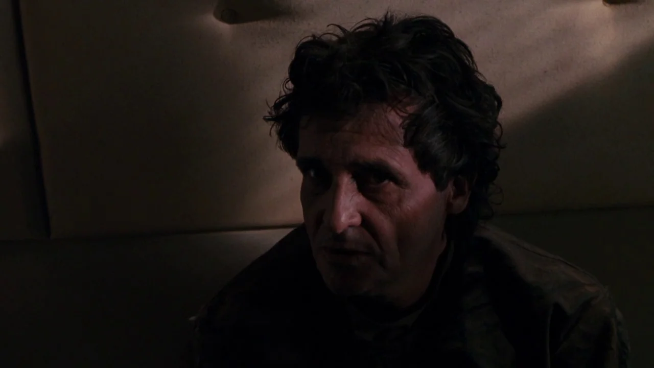 The Exorcist 3 (1990) movie still: closeup — A dimly lit interior space features padded surfaces and subtle shadows. A middle-aged man with dark,…
